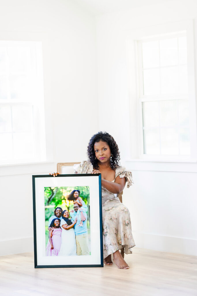 Photographer seated in a bright studio holding a large framed family portrait, showcasing custom wall art from a storytelling family photography session in Dallas.