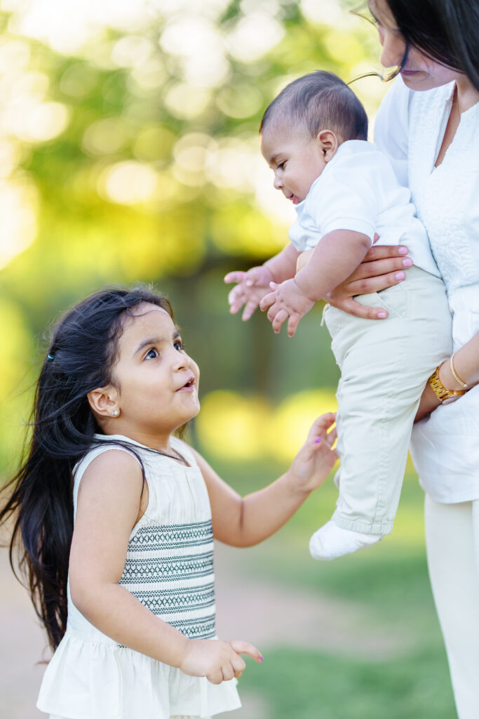 Little girl gently reaching for her baby sibling while mother holds the infant, a tender sibling moment during golden hour.