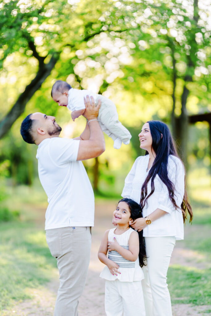 Father lifting baby into the air while mother and daughter smile nearby during a sunlit family moment in nature, capturing storytelling family photography in Dallas