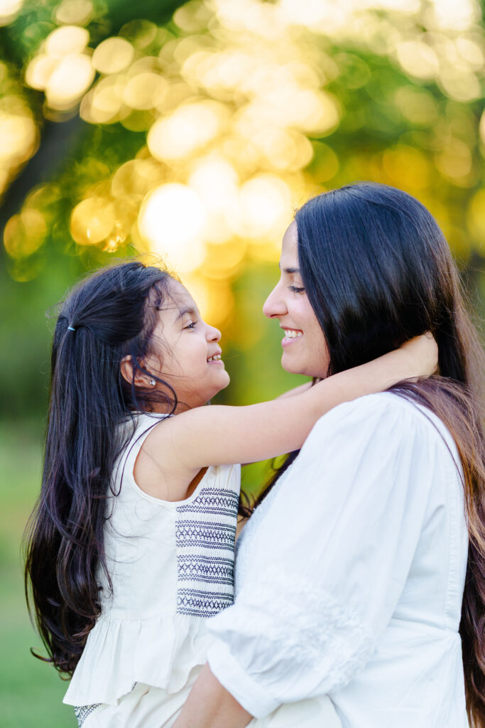 Close-up of daughter touching her mother’s face, both smiling with sunlit trees behind them—an intimate expression of love and presence.