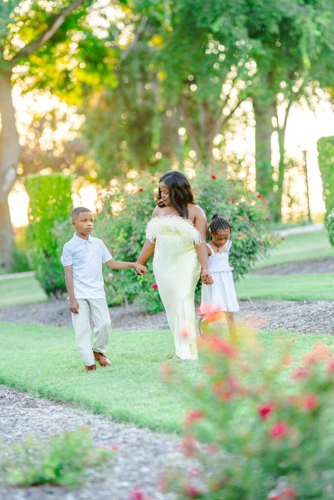 Frisco family photographer Tonaya Noel Photography captures a mother walking hand in hand with her children through blooming garden paths at sunset.
